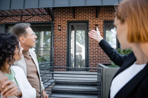 Agent immobilier flou dans des lunettes pointant avec la main sur la maison moderne près de couple multiethnique — Photo de stock