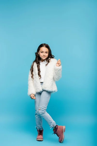 Полная длина позитивной дошкольницы в меховой куртке, позирующей на голубом фоне — стоковое фото