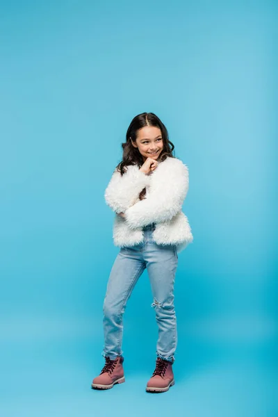 Longitud completa de niña preadolescente positiva en chaqueta de piel sintética y botas de invierno posando en azul - foto de stock