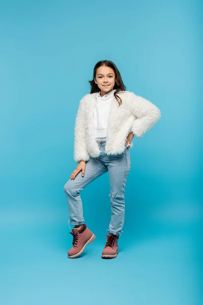 Comprimento total da menina pré-adolescente positiva em jaqueta de pele falsa e botas de inverno posando com a mão no quadril no azul — Fotografia de Stock