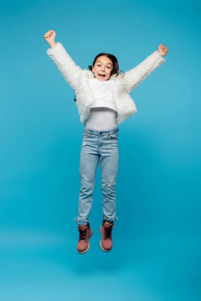 Full length of positive preteen girl in faux fur jacket and winter boots jumping on blue — Stock Photo
