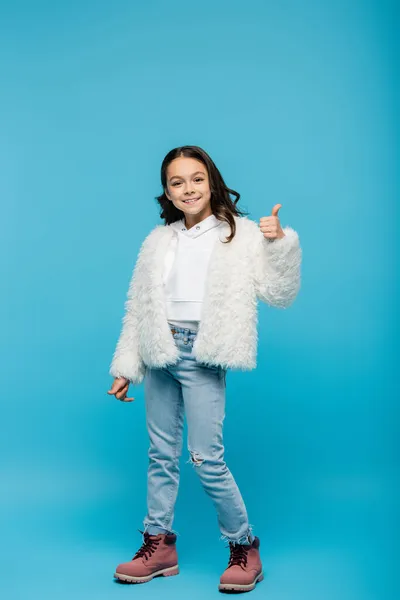 Full length of positive preteen girl in faux fur jacket and winter boots showing thumb up on blue — Stock Photo