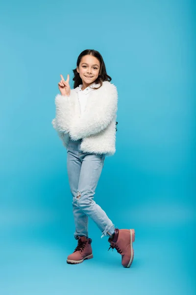 Full length of positive preteen girl in faux fur jacket and winter boots showing peace sign on blue — Stock Photo
