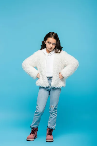 Full length of preteen girl in faux fur jacket and winter boots standing with hands on hips on blue — Stock Photo