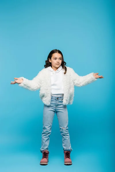 Longitud completa de la niña preadolescente en chaqueta de piel sintética y botas de invierno mostrando gesto de encogimiento de hombros en azul - foto de stock