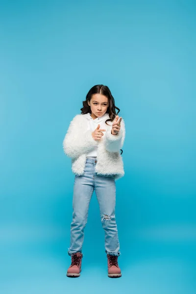 Piena lunghezza di preteen ragazza in giacca di pelliccia sintetica e stivali invernali mostrando gesto pistola dito su blu — Foto stock