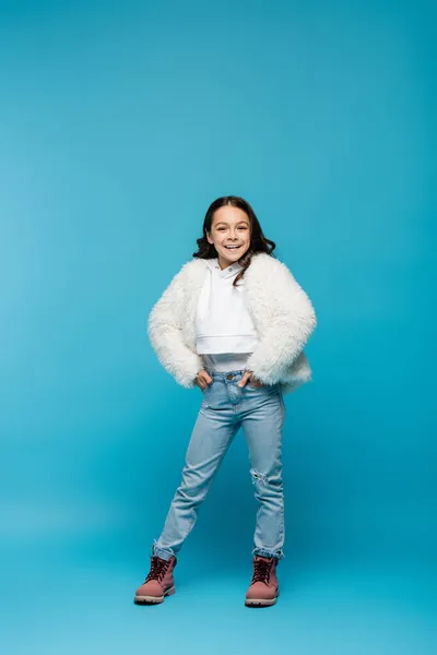 Longitud completa de la niña preadolescente feliz en chaqueta de piel sintética y botas de invierno de pie con las manos en los bolsillos en azul - foto de stock