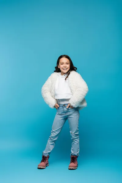 Pleine longueur de fille preteen heureux en fausse veste de fourrure et bottes d'hiver debout avec les mains dans les poches sur le bleu — Photo de stock