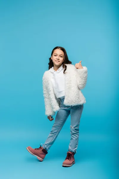 Повна довжина веселої дев'ятнадцяти дівчини в штучному піджаку і зимових чоботях, показуючи великий палець на синьому — стокове фото