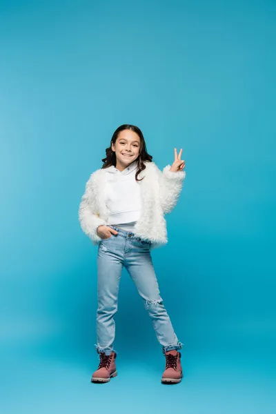 Full length of positive preteen girl in faux fur jacket and boots showing peace sign while posing with hand in pocket on blue — Stock Photo
