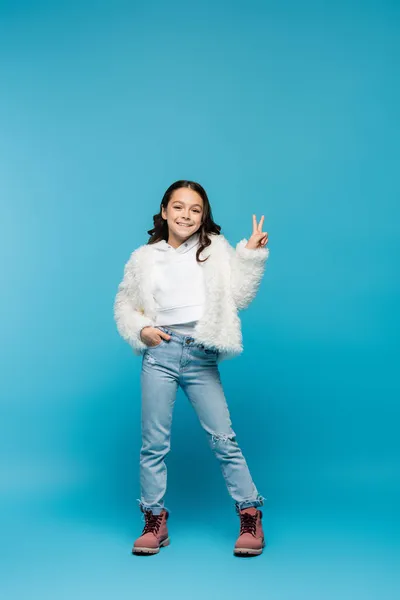 Comprimento total da menina pré-adolescente positiva em jaqueta de pele falsa e botas mostrando sinal de paz ao posar com a mão no bolso no azul — Fotografia de Stock