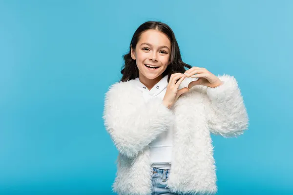 Chica preadolescente positiva en chaqueta de piel sintética blanca que muestra el corazón con las manos aisladas en azul - foto de stock