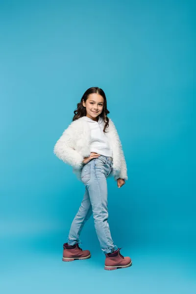 Повна довжина усміхненої дев'ятнадцяти дівчини в штучному піджаку і зимових чоботях, що стоять з рукою на стегні на синьому — стокове фото