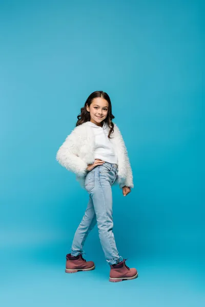 Pleine longueur de fille préadolescente souriante en fausse veste de fourrure et bottes d'hiver debout avec la main sur la hanche sur bleu — Photo de stock