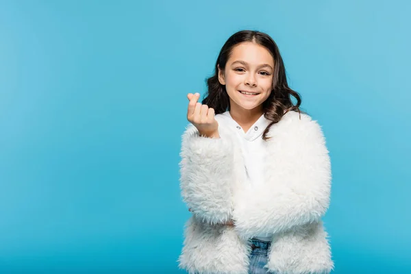 Feliz niña preadolescente en chaqueta de piel sintética blanca mirando a la cámara y mostrando el signo del corazón con los dedos aislados en azul - foto de stock