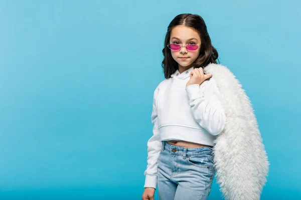 Preteen menina em óculos de sol rosa segurando jaqueta de pele branca falso isolado em azul — Fotografia de Stock