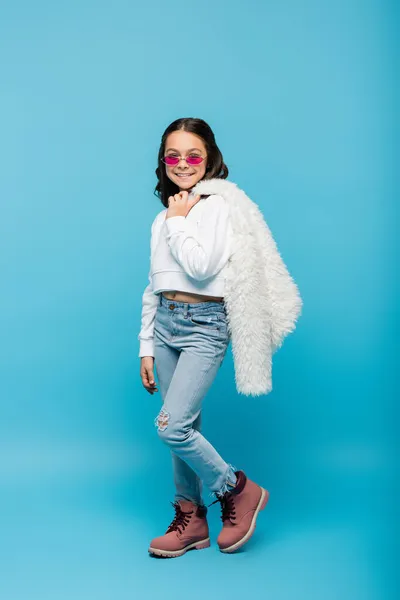 Comprimento total da menina pré-adolescente feliz em óculos de sol rosa segurando jaqueta de pele de falso branco no azul — Fotografia de Stock