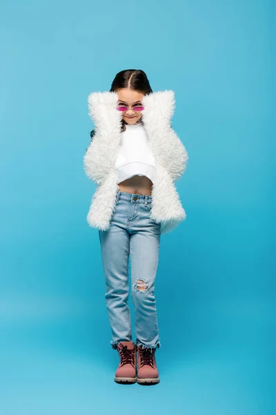 Full length of smiling preteen girl in pink sunglasses and white faux fur jacket on blue — Stock Photo