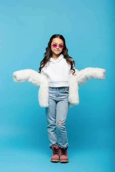 Full length of smiling preteen girl in pink sunglasses and white faux fur jacket showing shrug gesture on blue — Stock Photo