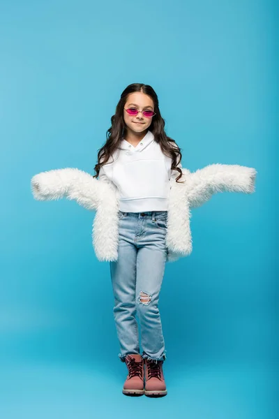 Pleine longueur de préadolescente souriante en lunettes de soleil roses et veste blanche en fausse fourrure montrant geste haussant les épaules sur bleu — Photo de stock