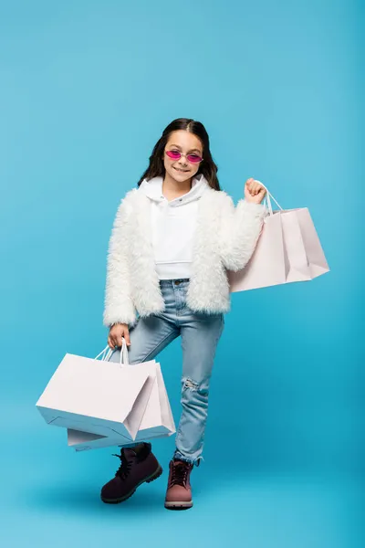 Pleine longueur de heureux preteen girl en lunettes de soleil roses et veste en fausse fourrure tenant des sacs à provisions sur bleu — Photo de stock