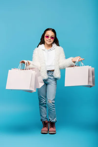 Повна довжина радісної дівчини в рожевих сонцезахисних окулярах і штучному піджаку, що тримає сумки на синьому — стокове фото