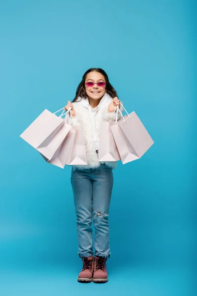 Full length of happy preteen girl in pink sunglasses and faux fur jacket holding shopping bags on blue — Stock Photo