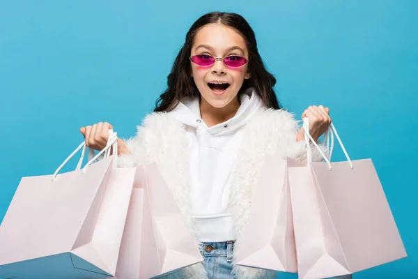 Stupito preteen girl in occhiali da sole rosa e giacca di pelliccia sintetica che tiene borse della spesa isolate su blu — Foto stock