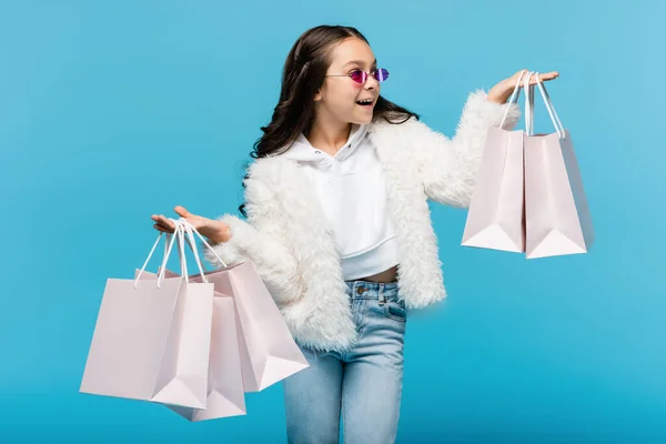 Animado preteen menina em óculos de sol rosa e casaco de pele falso segurando sacos de compras isolado no azul — Fotografia de Stock