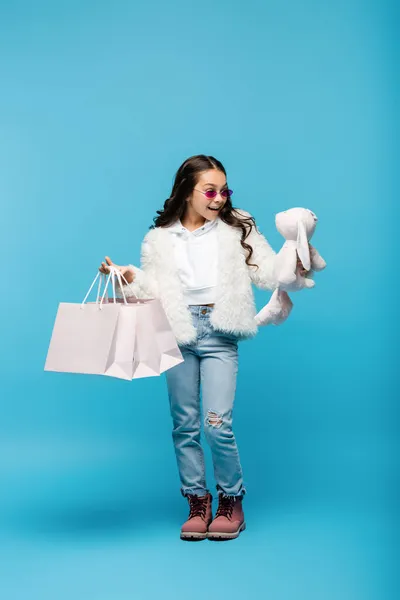 Longitud completa de la niña preadolescente feliz en gafas de sol de color rosa y chaqueta de piel sintética celebración de conejito suave y bolsas de compras en azul - foto de stock