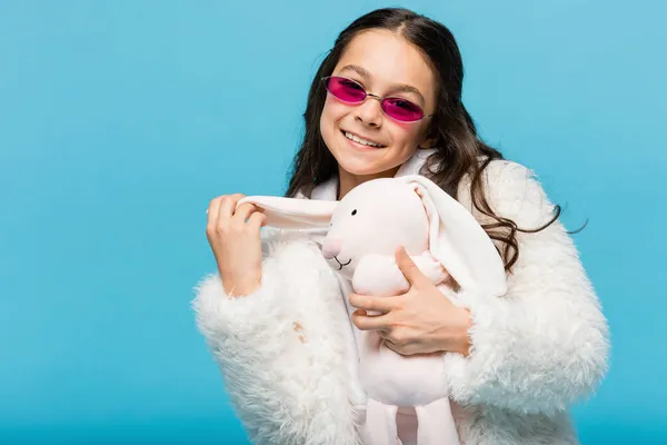 Happy preteen girl in pink sunglasses and faux fur jacket holding soft bunny isolated on blue — Stock Photo