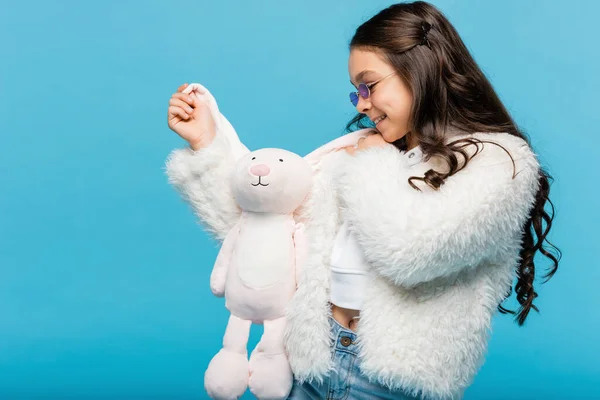 Menina pré-adolescente alegre em óculos de sol rosa e casaco de pele falso segurando coelho macio isolado em azul — Fotografia de Stock