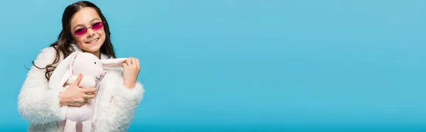 Felice preteen girl in occhiali da sole rosa e giacca di pelliccia sintetica tenendo morbido coniglietto isolato su blu, banner — Foto stock
