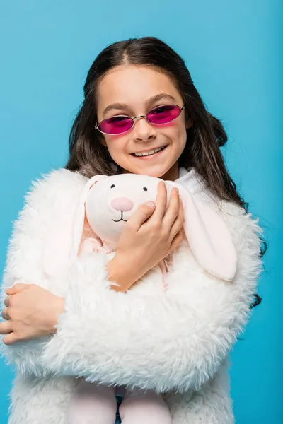 Cheerful preteen girl in pink sunglasses and faux fur jacket hugging soft bunny isolated on blue — Stock Photo