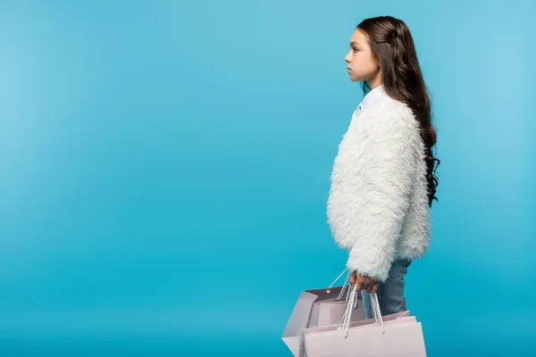 Seitenansicht eines Mädchens in weißer Kunstpelzjacke mit Einkaufstaschen auf blauem Grund — Stockfoto