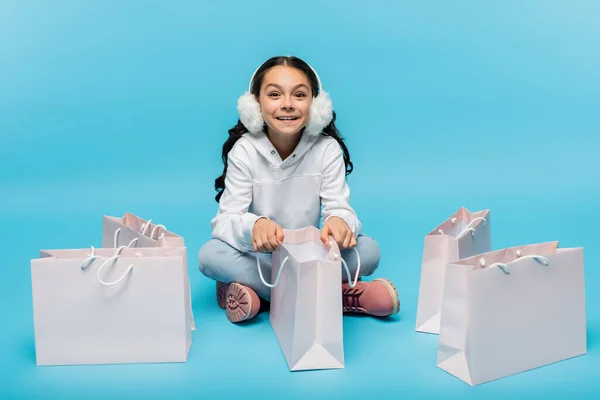 Longitud completa de alegre niña preadolescente en orejeras blancas y chaqueta de piel sintética sentado cerca de bolsas de compras en azul - foto de stock