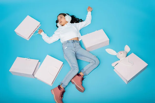 High angle view of tired preteen girl in ear muffs lying near shopping bags and soft bunny on blue — Stock Photo