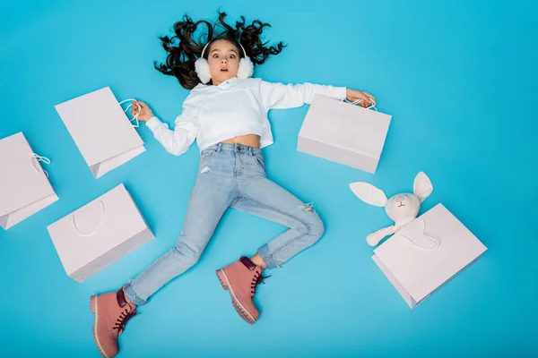 High angle view of shocked preteen girl in ear muffs lying near shopping bags and soft bunny on blue — Stock Photo