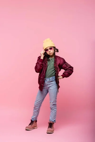 Volle Länge der preteen Mädchen im trendigen Outfit Einstellung Sonnenbrille und posiert mit der Hand auf der Hüfte auf rosa — Stockfoto