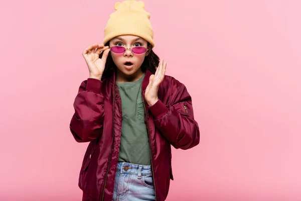 Impactada chica preadolescente en traje de moda ajustando gafas de sol aisladas en rosa - foto de stock