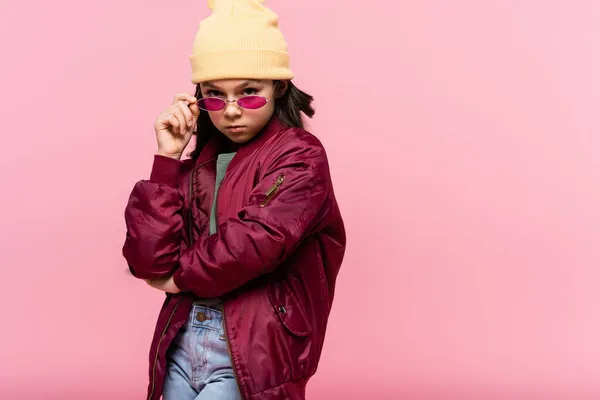 Preteen girl in trendy outfit and hat adjusting sunglasses isolated on pink — Stock Photo