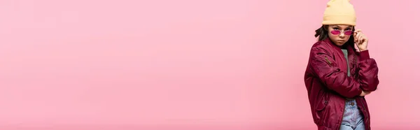 Preteen girl in abito alla moda e cappello regolazione occhiali da sole isolati su rosa, banner — Foto stock