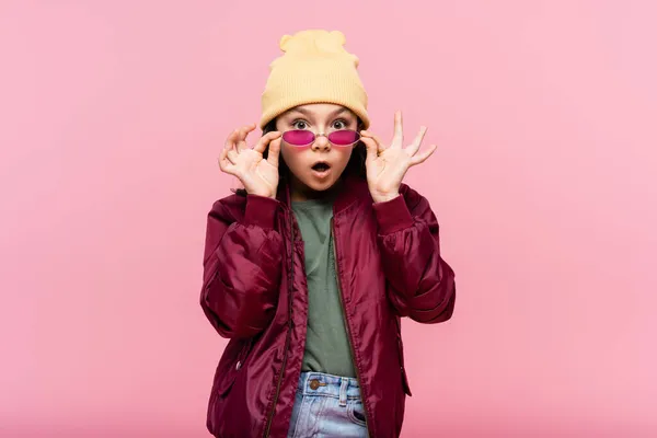 Surprised preteen girl in trendy outfit adjusting sunglasses isolated on pink — Stock Photo