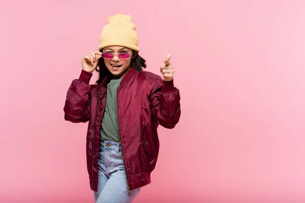 Happy preteen girl in trendy outfit adjusting sunglasses and pointing at camera isolated on pink — Stock Photo