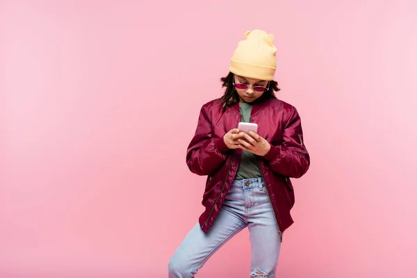 Niña preadolescente en traje de moda y gafas de sol usando teléfono inteligente aislado en rosa - foto de stock