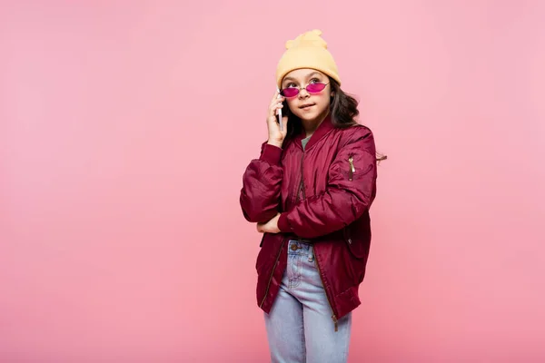Preteen girl in trendy outfit and sunglasses talking on smartphone isolated on pink — Stock Photo