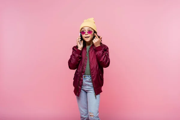 Niña preadolescente en traje elegante y gafas de sol hablando en el teléfono inteligente aislado en rosa - foto de stock