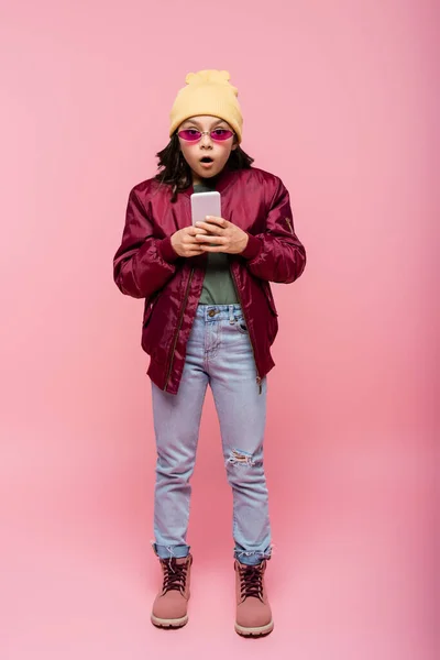 Piena lunghezza di scioccato preteen girl in abito alla moda utilizzando smartphone su rosa — Foto stock