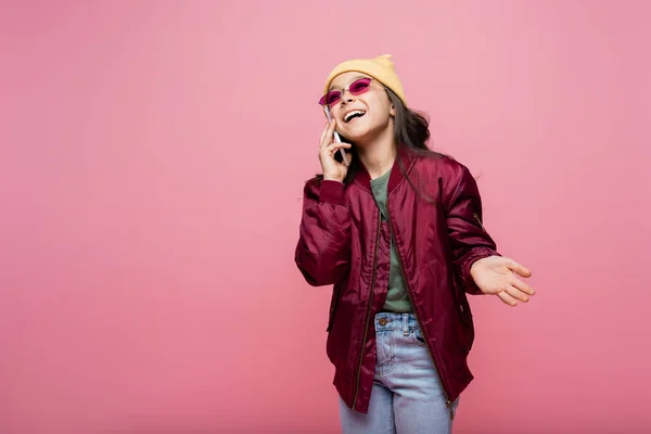 Happy preteen girl in stylish outfit and sunglasses talking on smartphone isolated on pink — Stock Photo