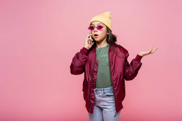Chocado preteen menina no elegante roupa e óculos de sol falando no smartphone enquanto gesticulando isolado no rosa — Fotografia de Stock